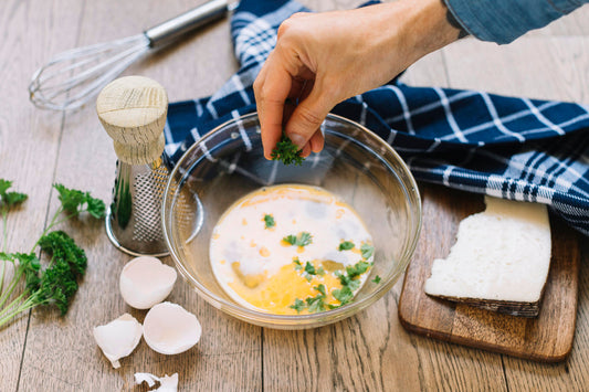 Hand adding fresh parsley to a bowl of eggs and oat milk on a wooden table, preparing scrambled eggs with herbs.