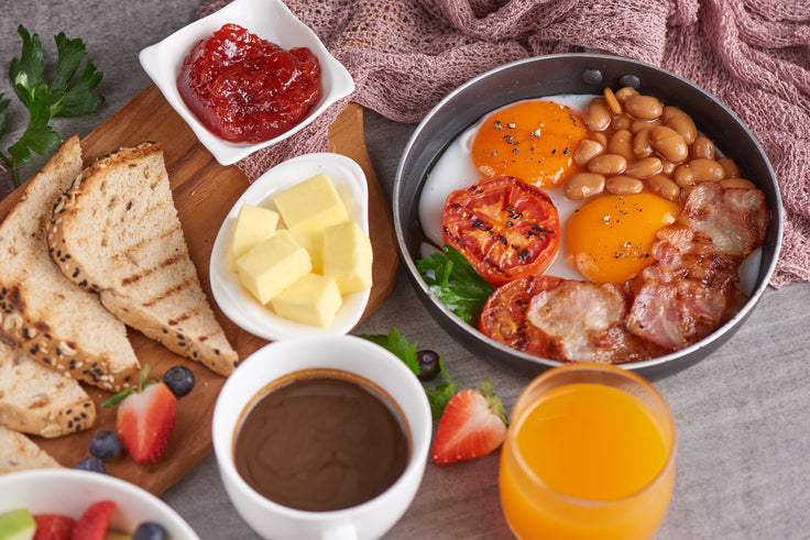 breakfast meal on top of the table with a pan of sunny side up eggs and beans and tomatoes, fresh orange juice, cup of chocolate drink, butter, sliced bread, and fruits