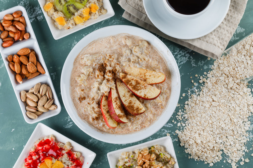 a bowl of oat meal with sliced apples on top beside a tray plate of nuts, fruits, and dried oats